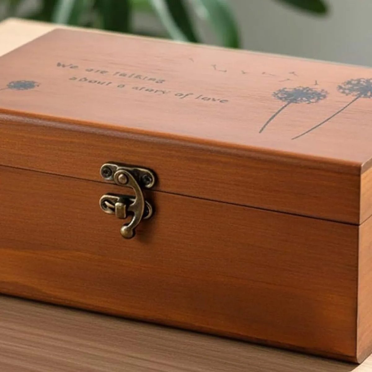 La caja de costura cerrada sobre una mesa de madera, con el broche de metal al frente y la tapa grabada con dos dientes de león